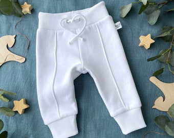 May include: White baby pants with a drawstring waist and ribbed cuffs. The pants have vertical seams down the front and are made of a soft material. The pants are displayed on a blue surface with wooden star decorations.