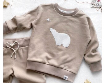 May include: A beige sweatshirt with a white embroidered polar bear and a star. The sweatshirt is paired with matching beige sweatpants.