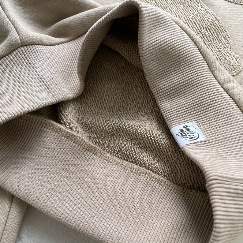 May include: A close-up of a beige sweatshirt with a ribbed collar and a small white label that says "smile's world".