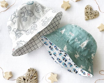 May include: Two reversible bucket hats. One hat is white with a blue bicycle print and a black and white checkered pattern on the reverse side. The other hat is teal with a white rabbit and flower print and a blue and white feather print on the reverse side.