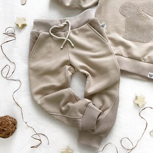 May include: Beige sweatpants with a drawstring waist and ribbed cuffs. The sweatpants have a contrasting panel on the side of each leg.