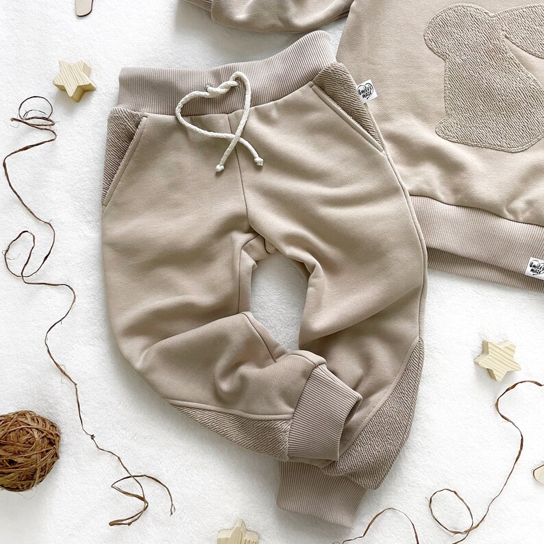 May include: Beige sweatpants with a drawstring waist and ribbed cuffs. The sweatpants have a contrasting panel on the side of each leg.