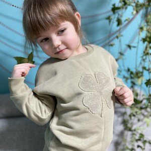 May include: A young child wearing a light green sweatshirt with a large embroidered four-leaf clover on the front. The sweatshirt has a crew neck and long sleeves.