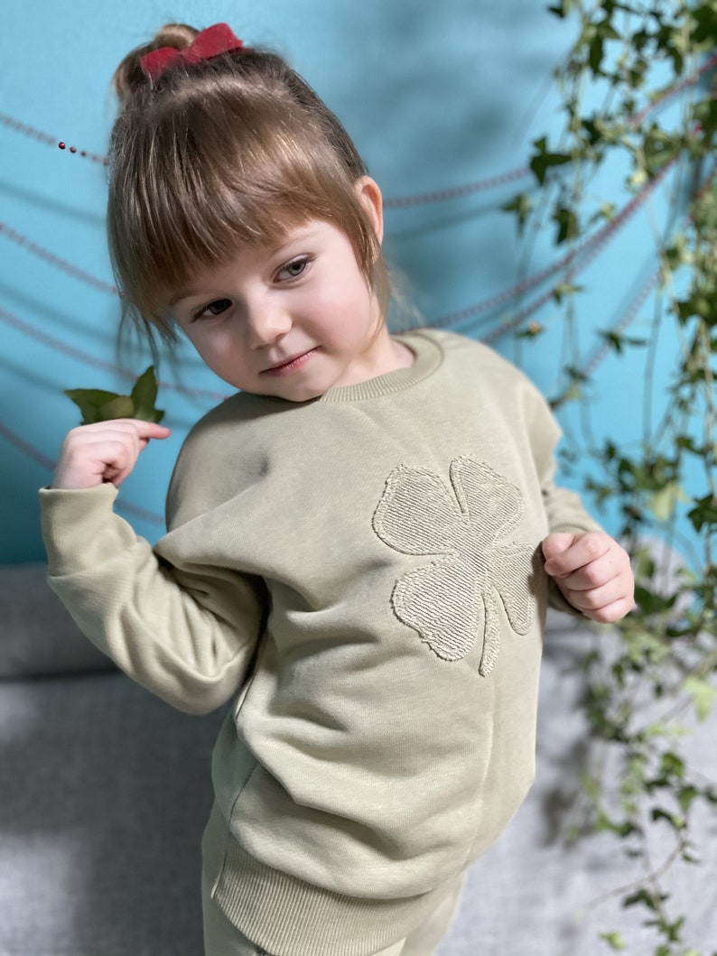 May include: A young child wearing a light green sweatshirt with a large embroidered four-leaf clover on the front. The sweatshirt has a crew neck and long sleeves.