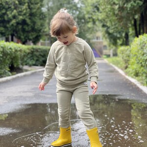 May include: A young child wearing a light green sweatshirt with a four-leaf clover design and matching sweatpants, standing in a puddle wearing yellow rain boots.