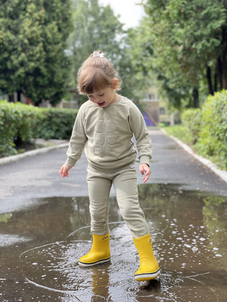 May include: A young child wearing a light green sweatshirt with a four-leaf clover design and matching sweatpants, standing in a puddle wearing yellow rain boots.
