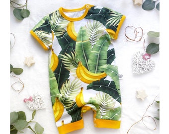 May include: A short-sleeved baby romper with a vibrant banana and tropical leaf print. The romper features a white background with yellow bananas and green leaves. It has yellow trim around the neck, sleeves, and legs.