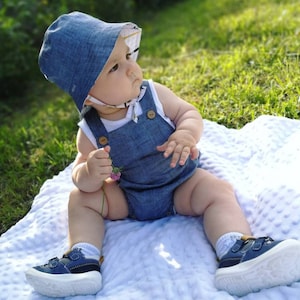 May include: A baby wearing a blue denim overall romper and a blue denim sun hat with a white and yellow patterned lining. The baby is sitting on a white blanket with a textured surface. The baby is holding a small pink flower.