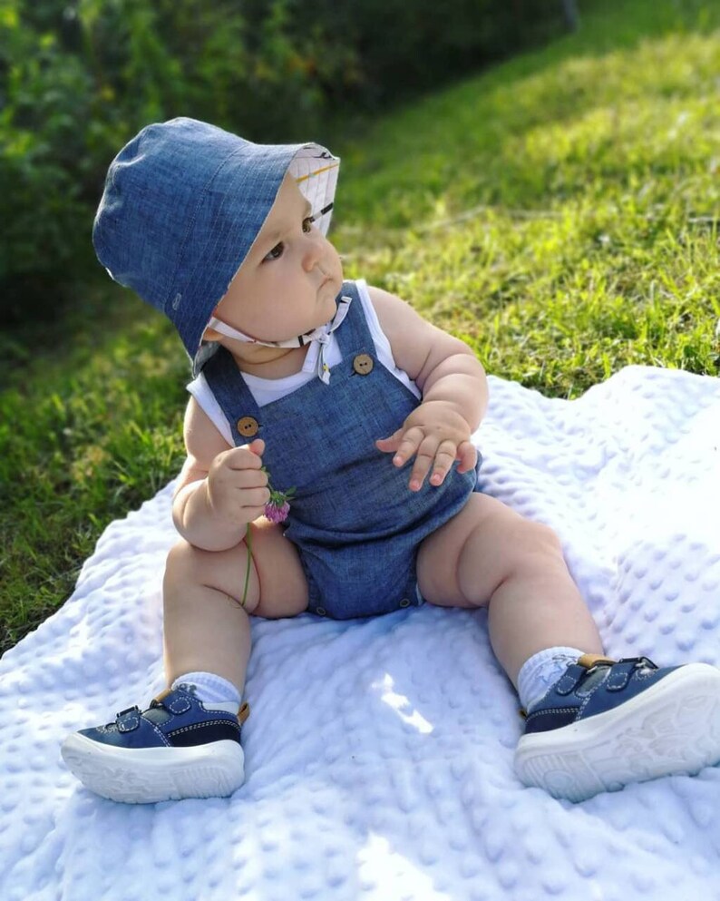 May include: A baby wearing a blue denim overall romper and a blue denim sun hat with a white and yellow patterned lining. The baby is sitting on a white blanket with a textured surface. The baby is holding a small pink flower.