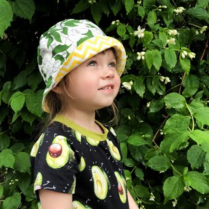 Może przedstawiać: Małe dziecko w białym i zielonym kwiatowym kapeluszu wiaderkowym z ż&oacute;łtym chevronem i czarnym t-shircie z zielonym nadrukiem awokado.