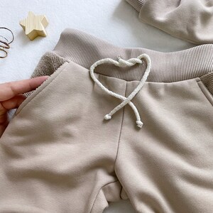 May include: Beige sweatpants with a white drawstring and a ribbed waistband. The sweatpants have a pocket on the side.