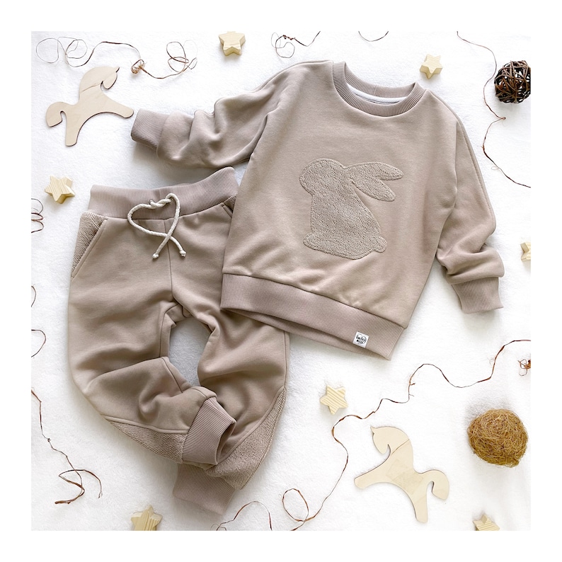 May include: A beige baby outfit with a bunny applique on the sweatshirt. The outfit includes a long-sleeved sweatshirt and matching sweatpants.