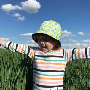 May include: A child wearing a striped long-sleeved shirt and a light blue bucket hat with a lemon print stands in a field of tall green grass.
