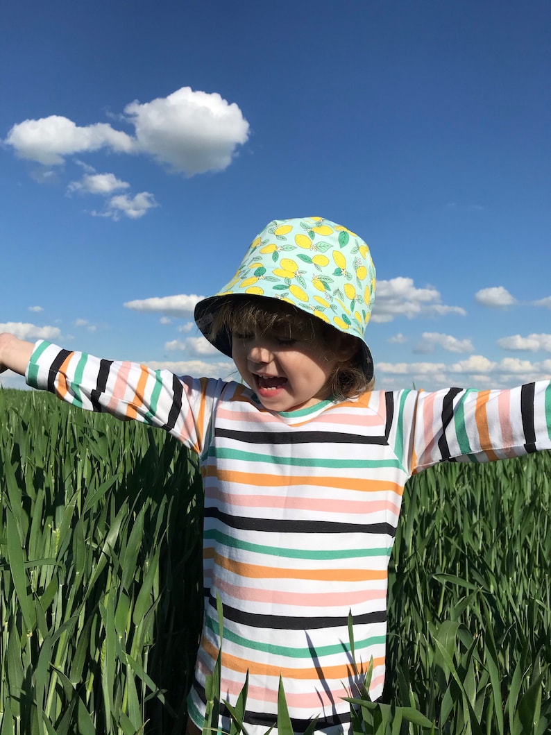May include: A child wearing a striped long-sleeved shirt and a light blue bucket hat with a lemon print stands in a field of tall green grass.