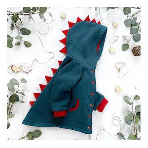 May include: A teal blue hooded jacket with a red dinosaur tail and red accents. The jacket has a front pocket and buttons.