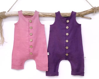 May include: Two sleeveless linen baby rompers, one in a dusty rose color and one in a deep purple color. Both rompers have a button closure down the front.