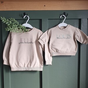 Könnte beinhalten: Zwei beige Sweatshirts hängen an weißen Kleiderbügeln. Auf dem linken Sweatshirt stehen der Name "ELLI" und die Worte "große Schwester". Auf dem rechten Sweatshirt stehen der Name "LOUIS" und die Worte "kleiner Bruder".