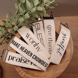 Könnte beinhalten: Cremefarbene Schlüsselanhänger aus Stoff mit schwarzer Schrift, darunter "Praise" und "Grateful". Jeder Schlüsselanhänger hat einen Metallverschluss. Die Schlüsselanhänger liegen in einer Holzschale, mit grünen Blättern im Hintergrund.