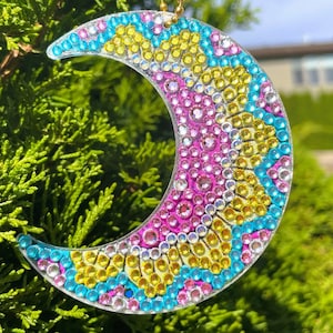 May include: A crescent moon-shaped ornament covered in colorful rhinestones. The ornament is made of clear plastic and features a pink, yellow, and blue design.