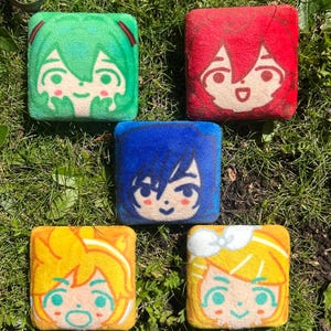 May include: Five plush, square-shaped pillows with anime-style character faces. The pillows are green, red, blue, yellow, and yellow with white. Each pillow features a different character design with detailed facial features and hair.