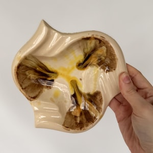 Peut inclure: Un plat en céramique beige de forme organique unique. L'intérieur présente un motif de teintes marron et jaune. Le plat est tenu sur un fond blanc, mettant en valeur son design et ses variations de couleurs. Une pièce décorative pour la maison.