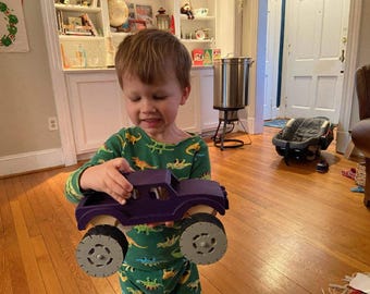 May include: A purple wooden toy truck featuring large, thick gray wheels with a textured tread pattern. The truck body has a simple, blocky design with an open cab area and a flatbed, showcasing a natural wood finish on the interior and wheel hubs.
