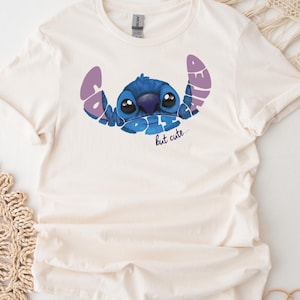 May include: Cream-colored t-shirt featuring a blue cartoon character with large eyes and the text "Bad but cute" in purple and blue. The shirt is made of a soft material and has short sleeves.