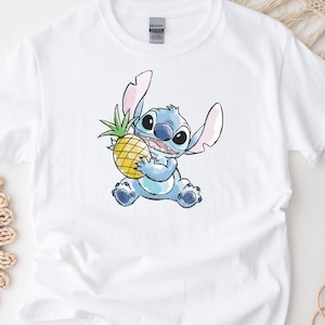K&ouml;nnte beinhalten: Wei&szlig;es T-Shirt mit einer Aquarell-Illustration von Stitch, einem blauen Alien-Charakter, der eine gelbe Ananas h&auml;lt. Das Design umfasst Stitchs gro&szlig;e Ohren und gro&szlig;en Augen. Das Shirt besteht aus einem weichen, wei&szlig;en Stoff.