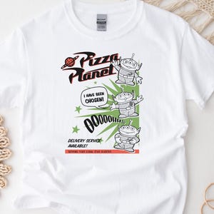 May include: White t-shirt featuring a graphic from Pizza Planet. The design includes the Pizza Planet logo, three green aliens, and the text "I HAVE BEEN CHOSEN!" and "DELIVERY SERVICE AVAILABLE!"