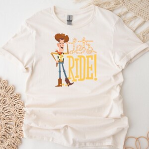 May include: Off-white t-shirt featuring a cartoon cowboy character with brown hair, a yellow sheriff's badge, and blue jeans. The text "Let's RIDE!" is printed in yellow. The shirt has short sleeves and a crew neck.