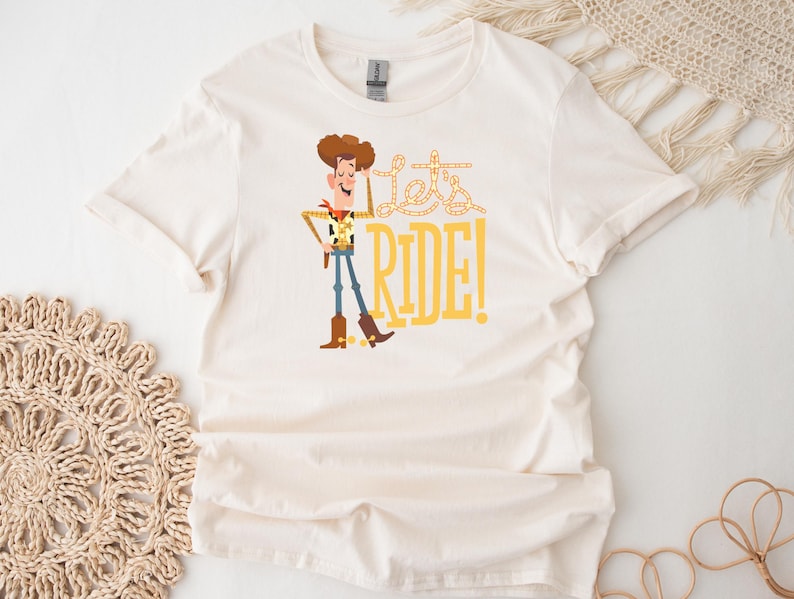 May include: Off-white t-shirt featuring a cartoon cowboy character with brown hair, a yellow sheriff's badge, and blue jeans. The text "Let's RIDE!" is printed in yellow. The shirt has short sleeves and a crew neck.