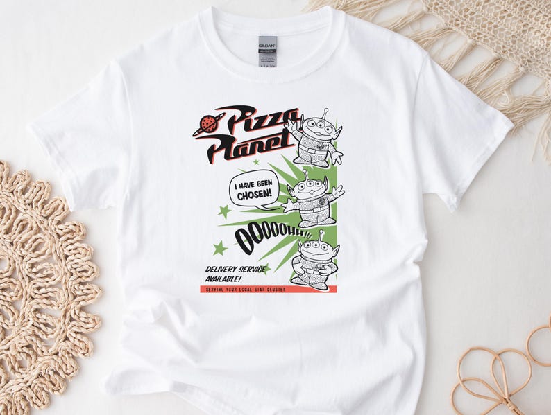 May include: White t-shirt featuring a graphic from Pizza Planet. The design includes the Pizza Planet logo, three green aliens, and the text "I HAVE BEEN CHOSEN!" and "DELIVERY SERVICE AVAILABLE!"