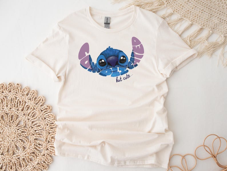 May include: Cream-colored t-shirt featuring a blue cartoon character with large eyes and the text "Bad but cute" in purple and blue. The shirt is made of a soft material and has short sleeves.