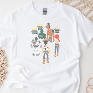 May include: White t-shirt featuring a colorful graphic of animated characters, including a cowboy, space ranger, and horse. The design also includes green aliens and a cowgirl. The shirt is made of a soft material.
