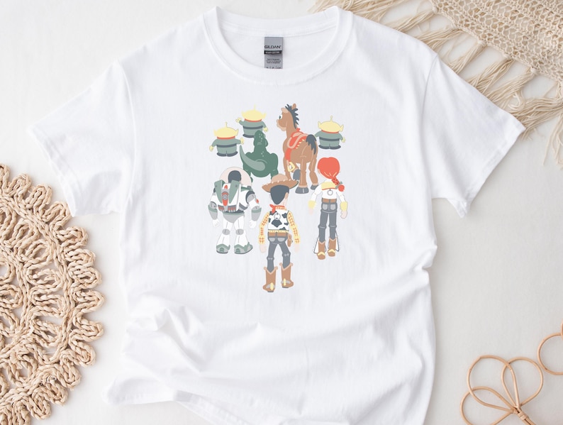 May include: White t-shirt featuring a colorful graphic of animated characters, including a cowboy, space ranger, and horse. The design also includes green aliens and a cowgirl. The shirt is made of a soft material.