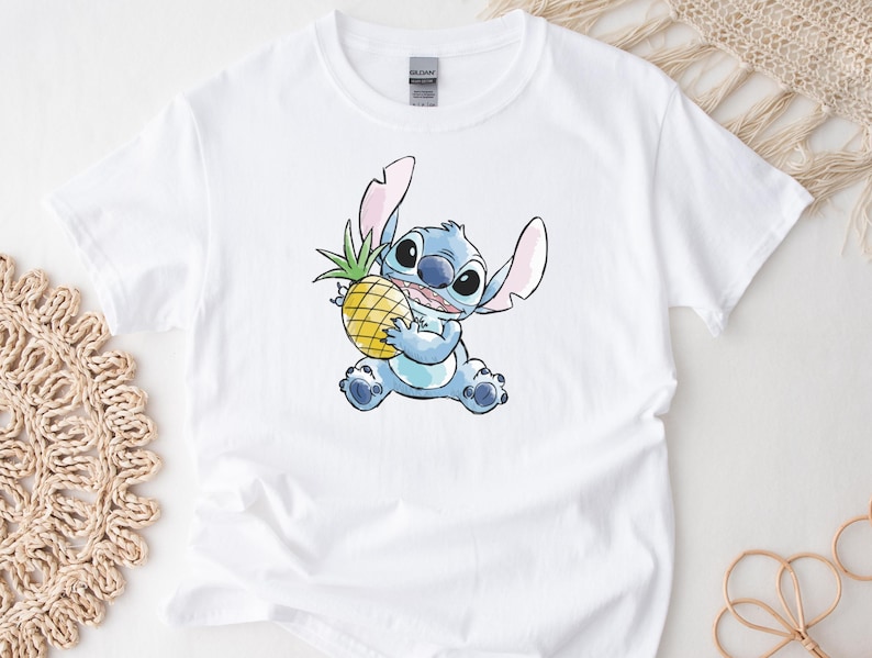 K&ouml;nnte beinhalten: Wei&szlig;es T-Shirt mit einer Aquarell-Illustration von Stitch, einem blauen Alien-Charakter, der eine gelbe Ananas h&auml;lt. Das Design umfasst Stitchs gro&szlig;e Ohren und gro&szlig;en Augen. Das Shirt besteht aus einem weichen, wei&szlig;en Stoff.