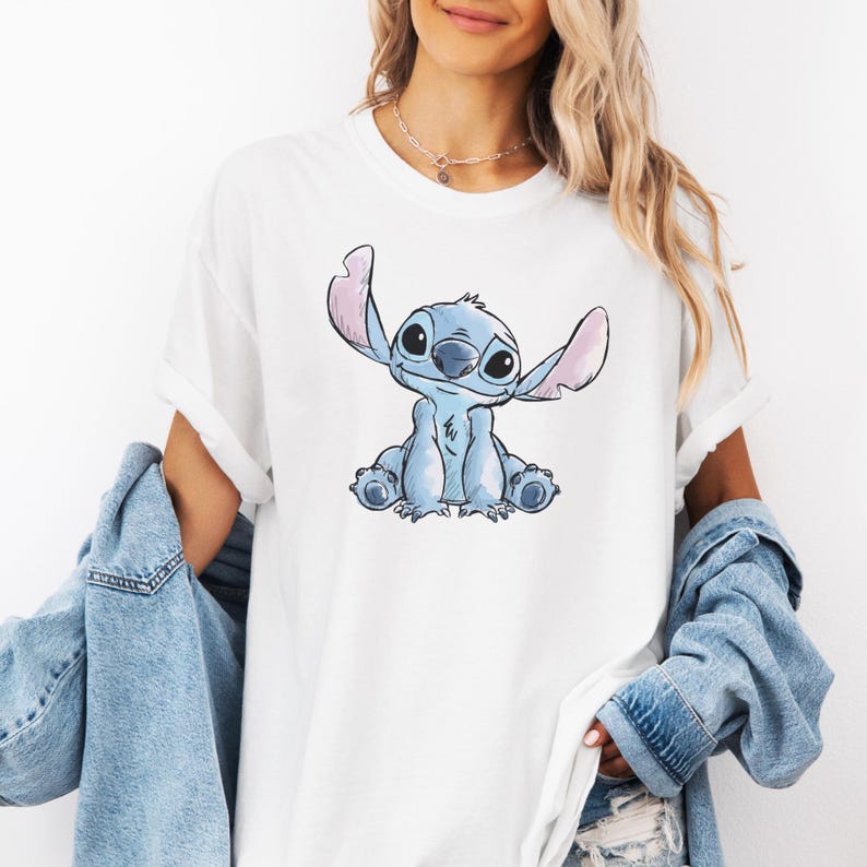 Op de afbeelding: Wit T-shirt met een blauw stripfiguur met grote oren en een vriendelijke uitdrukking. Het personage zit. Het shirt heeft opgerolde mouwen en wordt gecombineerd met een spijkerjasje.