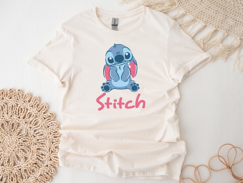 Puede incluir: Camiseta color crema con una ilustraci&oacute;n de dibujos animados de Stitch, un personaje alien&iacute;gena azul con grandes orejas y una expresi&oacute;n amigable. La palabra "Stitch" est&aacute; impresa en rosa debajo.