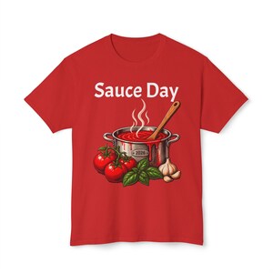 May include: A red t-shirt featuring a graphic of a silver pot filled with red tomato sauce, a wooden spoon, three red tomatoes, green basil leaves, and a head of garlic. The text Sauce Day is printed in white above the pot, with 2026 on the pot.
