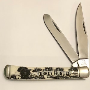May include: A pocket knife with two open stainless steel blades. The handle is off-white with a black illustration of a turkey and the words "TURKEY HUNTER". The knife has silver-colored metal accents on each end.