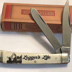May include: A stainless steel pocket knife with a cream-colored handle featuring a black illustration and the text "Logger's Life." The knife is partially open, revealing the sharp blades. The box behind the knife has the brand name "ROUGH RYDER."