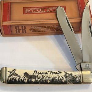 May include: A folding pocket knife with two silver blades and a cream-colored handle featuring a black graphic of a pheasant, a hunting dog, and a person with a rifle, with the text Pheasant Hunter. The knife is displayed before its box.