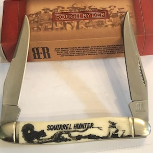 May include: A Rough Ryder Squirrel Hunter pocket knife with two open blades. The handle is off-white with a black illustration of a squirrel and a dog. The knife is displayed in front of its original box.
