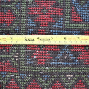Op de afbeelding: Close-up van een zwart, rood en blauw gepatroneerd tapijt met een gele meetlint eroverheen. De tape geeft "inch" en "cm" aan en is gemarkeerd tot 24 inch.