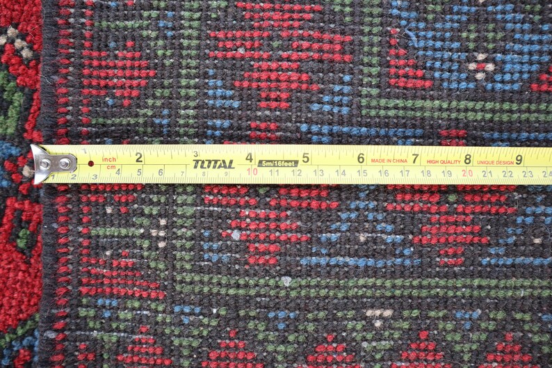 Op de afbeelding: Close-up van een zwart, rood en blauw gepatroneerd tapijt met een gele meetlint eroverheen. De tape geeft "inch" en "cm" aan en is gemarkeerd tot 24 inch.