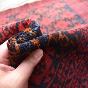 May include: Close-up of a red, black, and orange patterned rug with a fringe border. The rug is folded in half and being held by a hand.