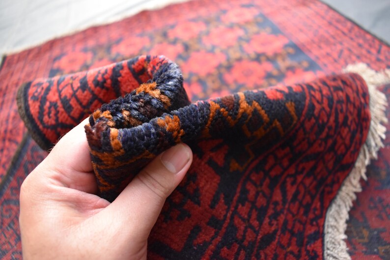May include: Close-up of a red, black, and orange patterned rug with a fringe border. The rug is folded in half and being held by a hand.