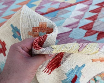 May include: A close-up of a hand holding a woven rug with a geometric pattern in shades of red, blue, yellow, and pink.