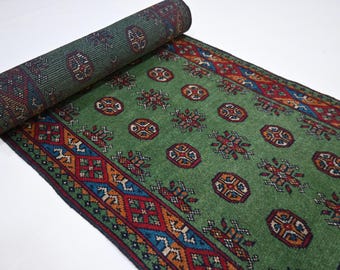May include: A rolled-up green rug with a detailed pattern. The rug features a central field of green with repeating circular and floral motifs in red, orange, and brown. The border has geometric designs in blue, red, and orange.
