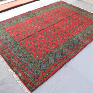 Op de afbeelding: Een rood tapijt met een geometrisch patroon in tinten van groen en blauw. Het tapijt heeft een franje rand.